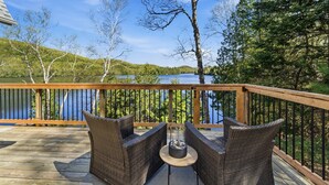 A wooden deck with wicker chairs and a table, overlooking a lake with trees and hills in the background.