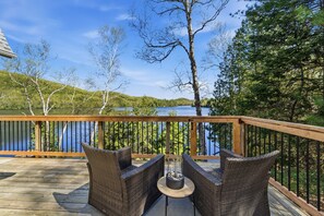 A wooden deck with wicker chairs and a table, overlooking a lake with trees and hills in the background.