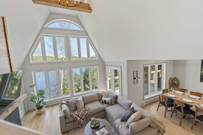 A spacious loft with a large window, a cozy sectional sofa, a dining table set for four, and a television in the corner.