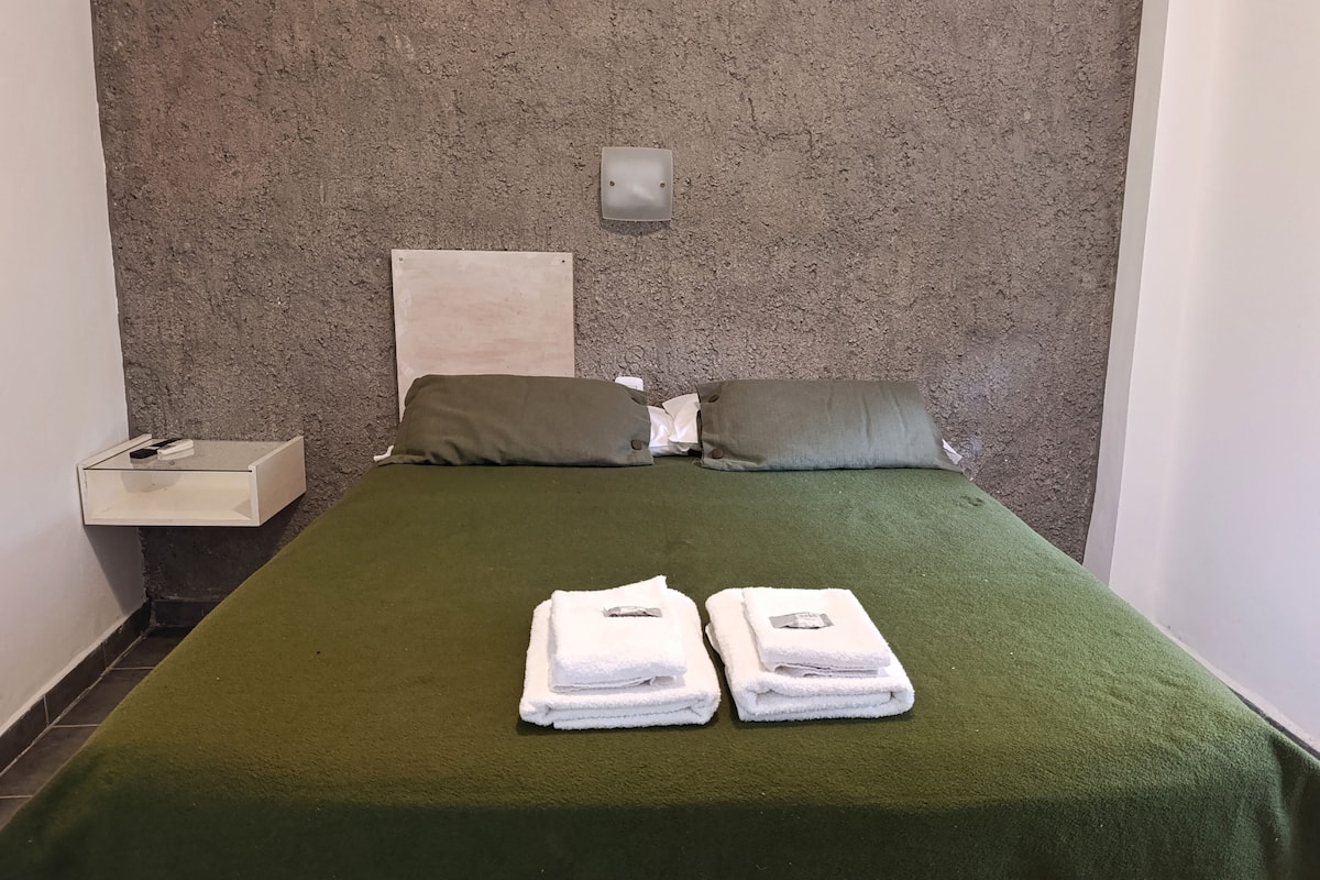 A room with a bed, two pillows, a nightstand, and a wall-mounted light fixture.