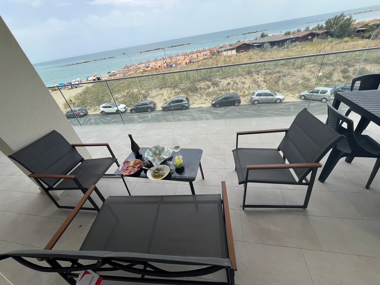 A balcony with two chairs, a table, and a bottle of wine. The view includes a beach and parked cars.