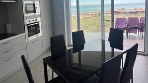 Ett modernt kök med inbyggda vitvaror, en matplats med ett glasbord och stolar samt utsikt över havet genom stora skjutdörrar.