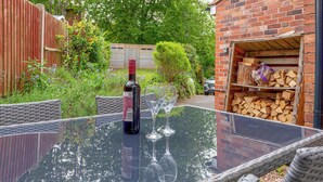 Une table avec un plateau en verre, une bouteille et deux verres à vin, une chaise en osier et un mur en briques avec une porte de garage.