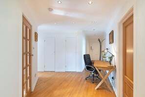 Ein Zimmer mit Holzboden, einem Schreibtisch mit Stuhl, einer Lampe und einem Blumentopf. Weiße Türen und ein Holztürrahmen sind zu sehen.