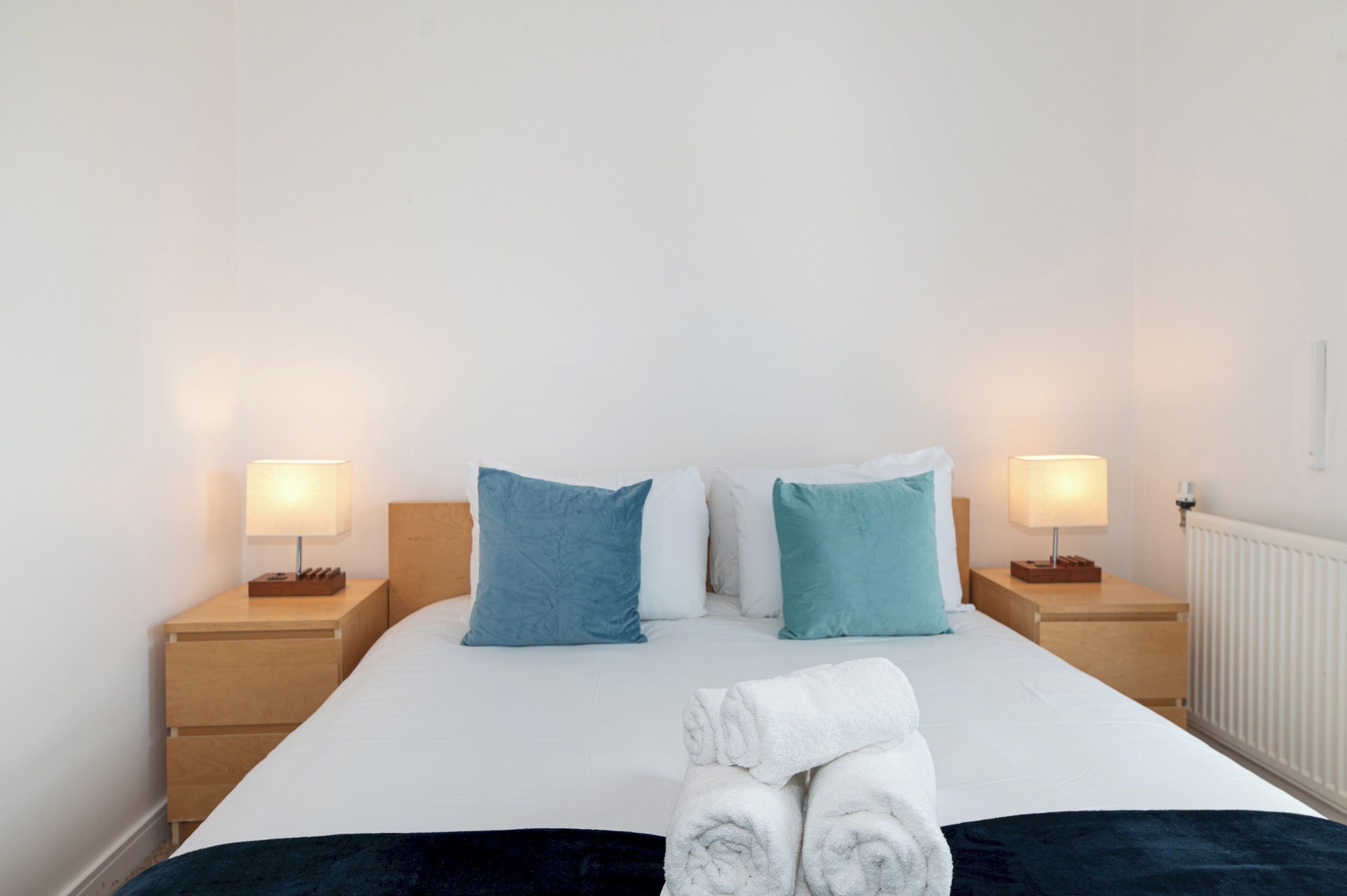 Una cama bien hecha con sábanas blancas, dos almohadas azules y un par de toallas blancas enrolladas sobre la cama. Hay dos mesitas de noche con lámparas, y un radiador blanco en la pared.