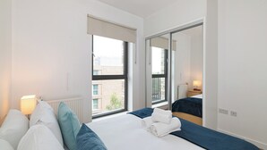 A hotel room with a large bed, a window with a view of buildings, a radiator, and a bedside table with a lamp. - Modern 2BR flat with Balcony, in East London - Pass the Keys (London)