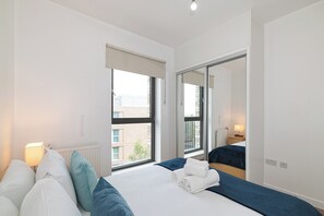 Ein Hotelzimmer mit einem großen Bett, einem Fenster mit Blick auf Gebäude, einem Heizkörper und einem Nachttisch mit Lampe.