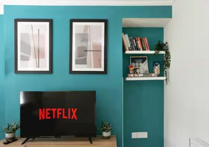 Ein Wohnzimmer mit Fernseher, der 'NETFLIX' anzeigt, abstrakten Kunstwerken an der Wand und einem Bücherregal mit Büchern und Dekorationsobjekten.