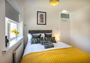 Ein ordentlich bezogenes Bett mit einer gelben Decke, einem gerahmten Kunstwerk darüber und einem Fenster mit Blumen auf dem Sims.