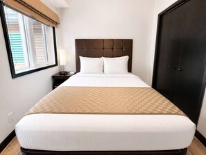 Ein Hotelzimmer mit einem großen Bett, einem Nachttisch, einer Lampe, einem Fenster mit Jalousien und einem Kleiderschrank.