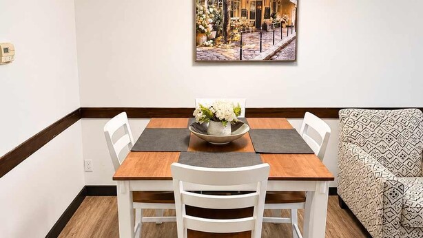 Zona pranzo con tavolo in legno, sedie bianche, un vaso con fiori e un quadro raffigurante un caffè al muro.
