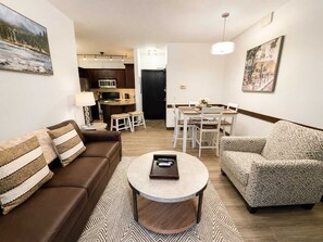 Ein Wohnzimmer mit braunem Sofa, gemustertem Sessel und Couchtisch. Es gibt eine Essecke mit Tisch und Stühlen, und im Hintergrund eine Küche.