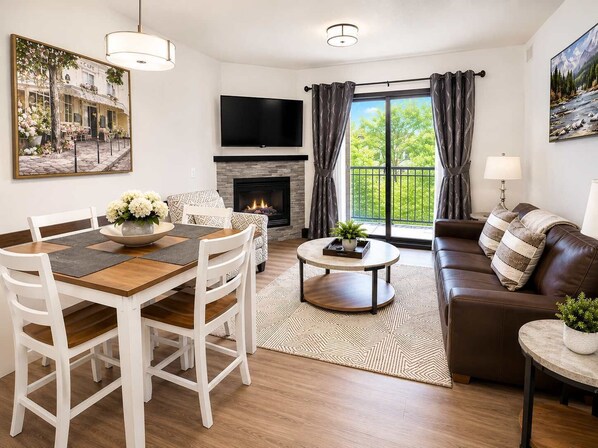 Ein Wohnzimmer mit Kamin, einem Esstisch mit Stühlen, einer Couch, einem an der Wand montierten Fernseher und einem großen Fenster mit Vorhängen.