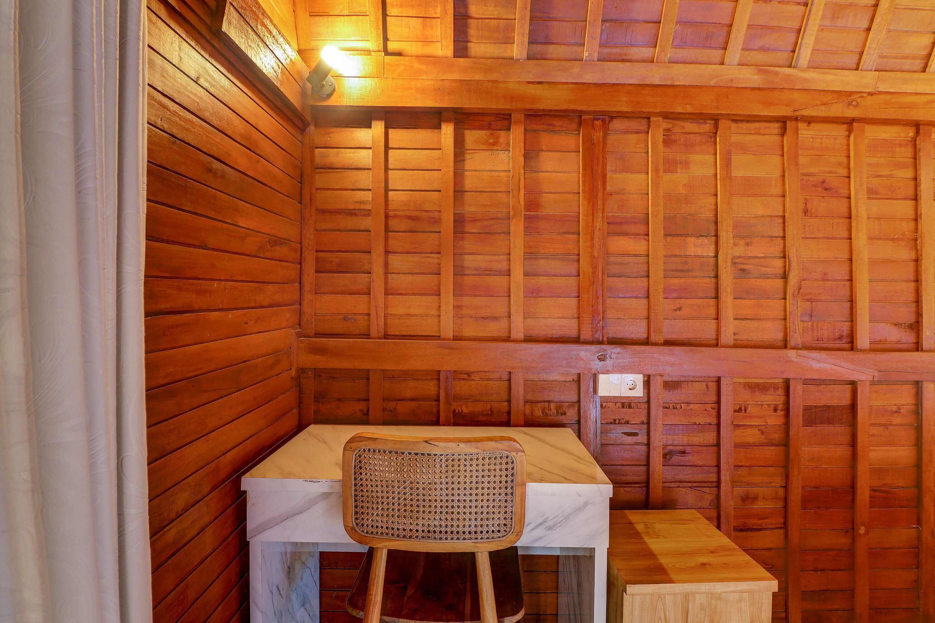 Interior de uma cabana de madeira com uma mesa, uma cadeira, um banco de madeira e um candeeiro fixado na parede.