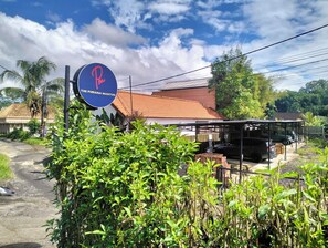 A sign for 'The Puriana Mantun' with a red and blue logo, surrounded by greenery and a building with a tiled roof.