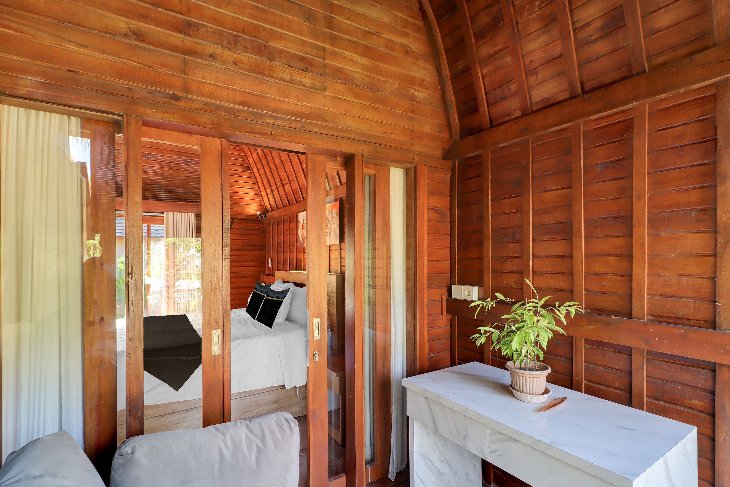 A wooden cabin interior with a bed, a small table with a plant, and a window.