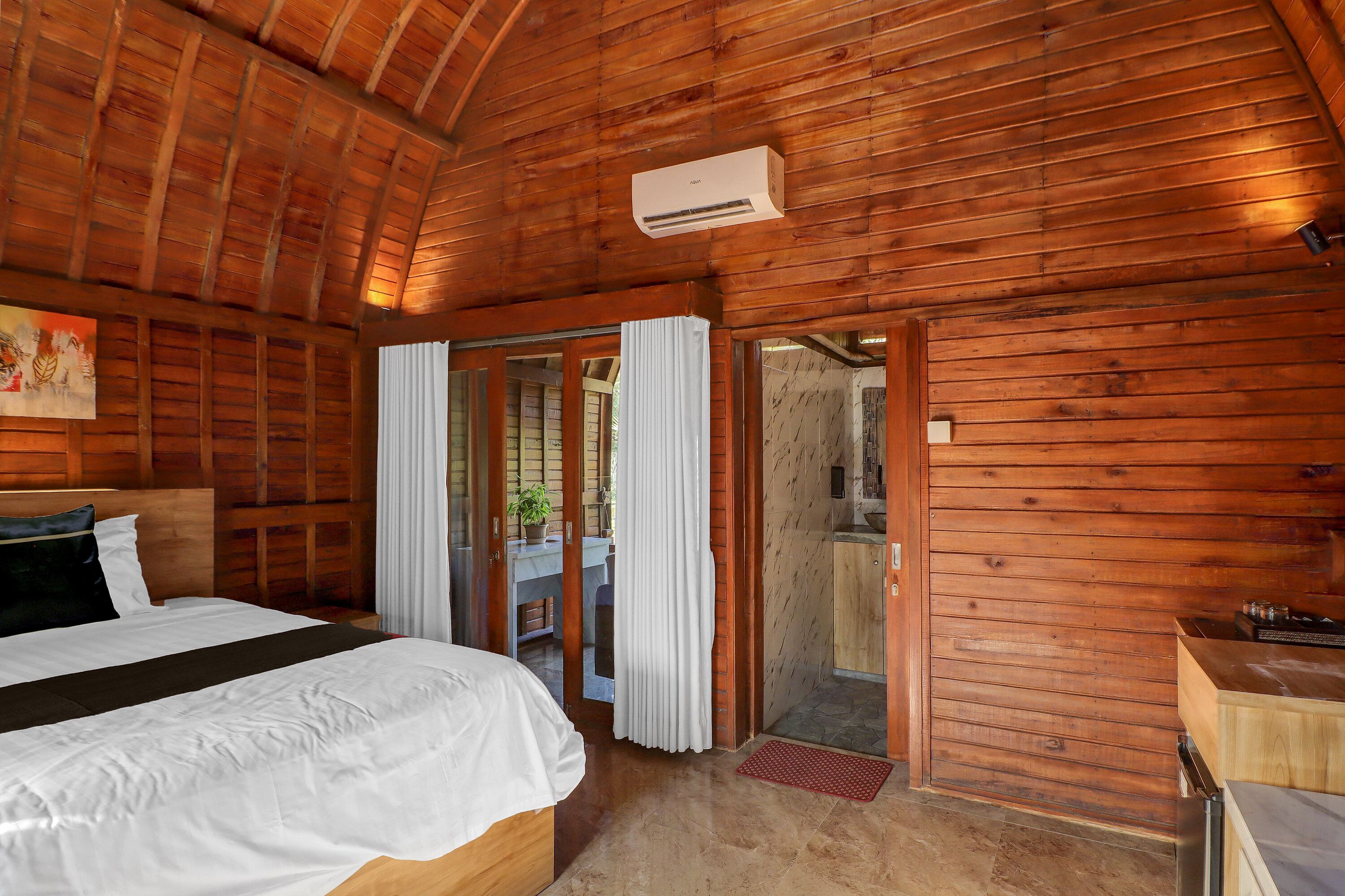 A wooden room with a bed, a small kitchenette, and a sliding glass door leading to an outdoor area.