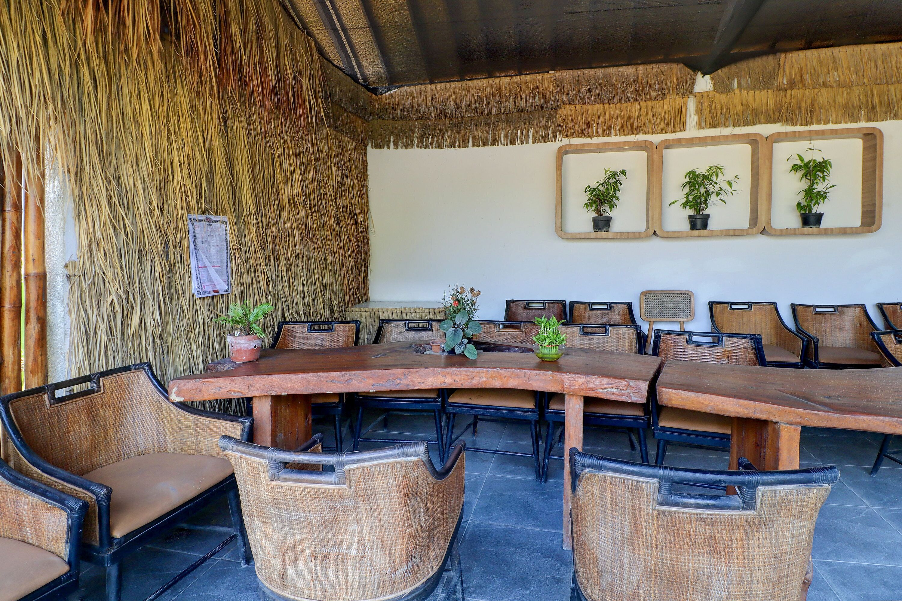 A covered outdoor dining area with a long wooden table surrounded by wicker chairs, potted plants, and framed pictures on the wall.