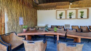 A covered outdoor dining area with a long wooden table surrounded by wicker chairs, potted plants, and framed pictures on the wall.
