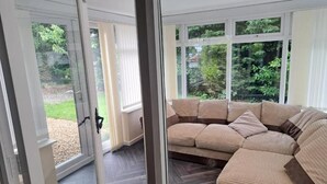 Moderni olohuone, jossa on suuri kulmasohva, lasiovi, joka johtaa ulos, ja näkymä vehreään maisemaan.