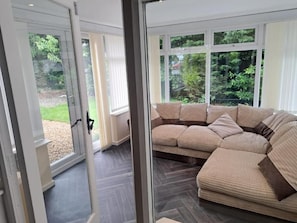 A modern living room with a large sectional sofa, a glass door leading outside, and a view of greenery.