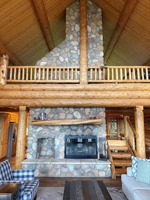 A log cabin interior with a stone fireplace, wooden staircase, and checkered sofas.