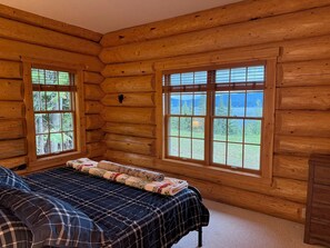 A log cabin bedroom with a bed, two windows, and a view of a forest and lake.