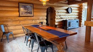 En inredning i en stuga byggd av trästockar, med ett matbord, stolar, en hängande lampa, en inramad tavla och en klocka på väggen.