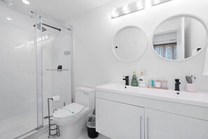 Un baño moderno con cabina de ducha de vidrio, inodoro, lavabo con artículos de aseo y dos espejos redondos.