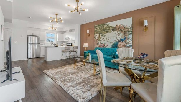 A modern living room with a teal sofa, a glass dining table, and a kitchen in the background. - Stylish Penthouse with Rooftop & Parking Included! (Niagara Falls)