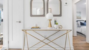 A white console table with gold accents, two mirrors, a white lamp, and a potted plant. - New 2BR Suite Sleeps 5, 7 Min to Falls w/Parking (Niagara Falls)