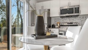 A modern kitchen with stainless steel appliances, a dining area with a glass table and white chairs, and a view of the outdoors through large windows. - New 2BR Suite Sleeps 5, 7 Min to Falls w/Parking (Niagara Falls)