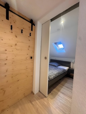 Une chambre avec un mur en lambris de bois, une lucarne, un lit avec une literie à motifs, une table de chevet et une commode.