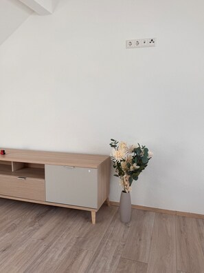 Ein schlichter Raum mit einem Holzschrank, einer Vase mit Blumen und einer weißen Wand mit Steckdosen.