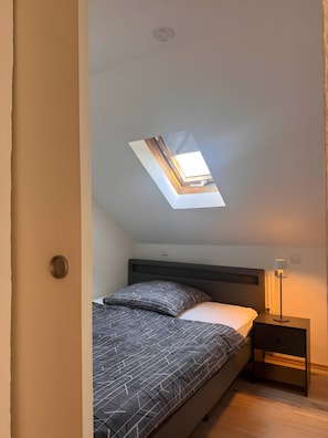 Une chambre avec un puits de lumière, un lit avec une literie à motifs, une table de chevet avec une lampe et un plancher en bois.