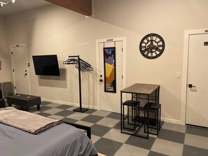 A hotel room with a bed, a TV, a desk with chairs, a dartboard, and a door with a colorful poster.