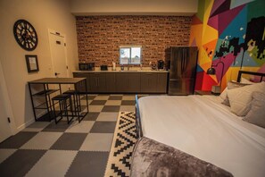 Een slaapkamer met een bed, een bureau met twee stoelen, een keukenhoek en een bakstenen muur.