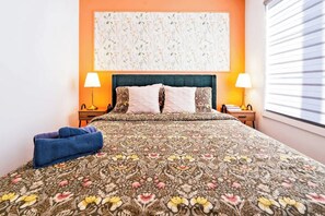 A bed with floral bedding, two bedside lamps, a blue towel, and a wall with a floral pattern. - Patio Suite with BBQ & Outdoor Relaxation, Near Niagara Falls! (Niagara Falls)