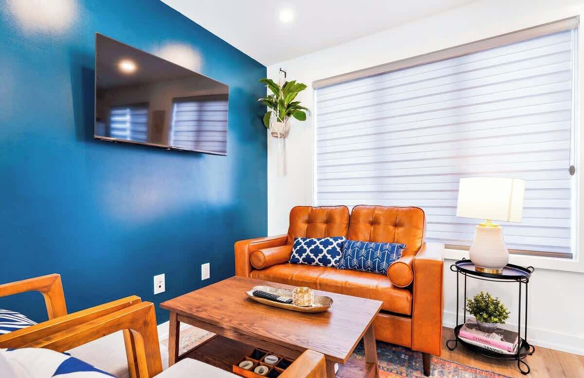 Un salon avec un mur bleu, un canapé en cuir, une table basse, un téléviseur fixé au mur et une fenêtre avec des stores.
