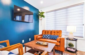 A living room with a blue wall, a leather sofa, a coffee table, a TV mounted on the wall, and a window with blinds. - Patio Suite with BBQ & Outdoor Relaxation, Near Niagara Falls! (Niagara Falls)