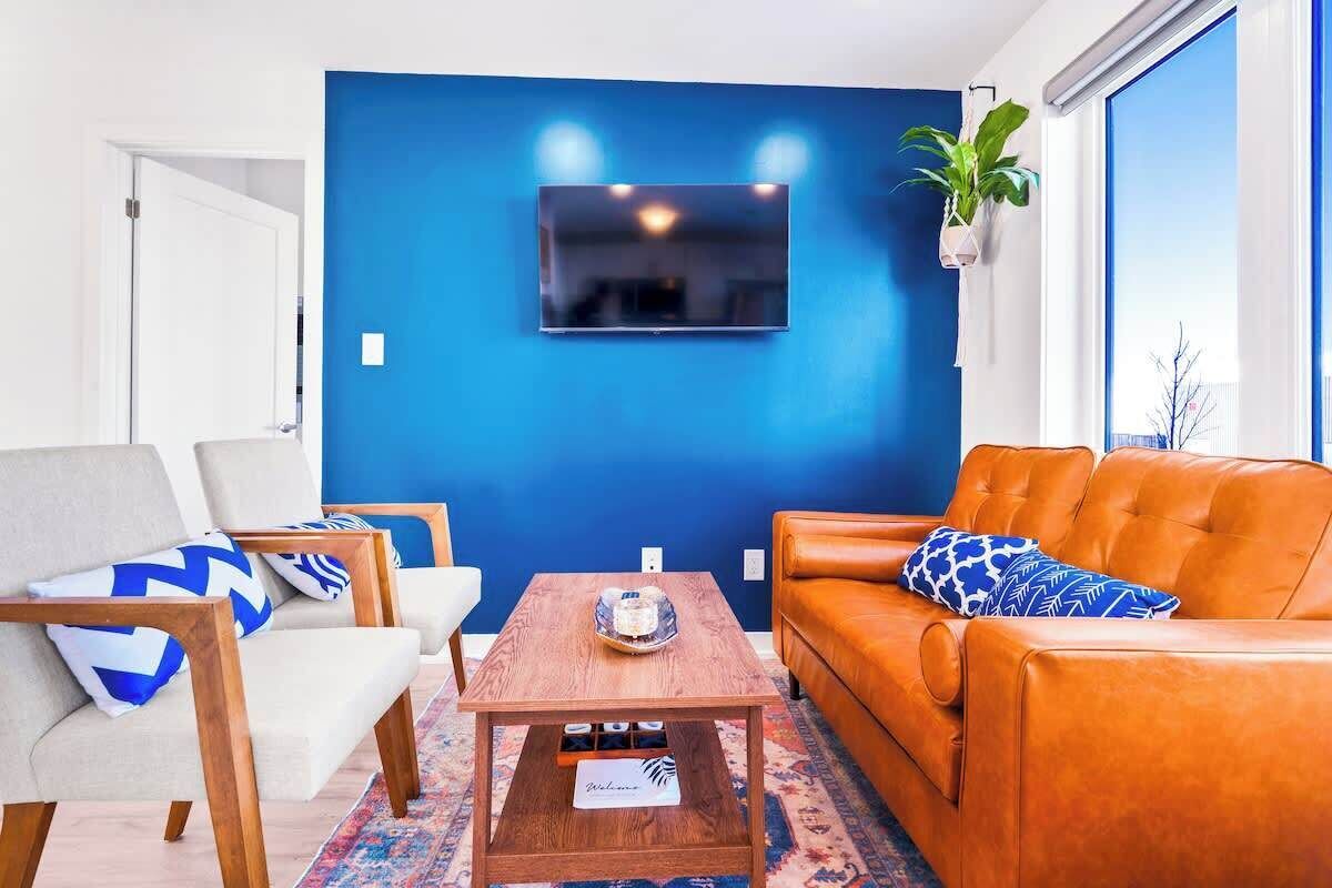Een woonkamer met een blauwe wand, een tv boven een houten salontafel, een leren bank met gestreepte kussens en een potplant.