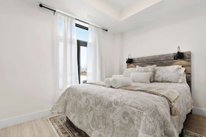 A bedroom with a large bed, a wooden headboard, a window with sheer curtains, and a view of the outdoors. - Spacious 2-Bedroom Suite, Mins to the Falls, Parking & Sleeps 5! (Niagara Falls)
