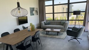 Una sala de estar moderna con un sofá, una mesa de comedor con sillas, una mesa de centro, un televisor montado en la pared y amplios ventanales con vistas al exterior.