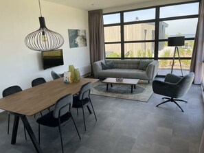 Een moderne woonkamer met een bank, een eettafel met stoelen, een salontafel, een aan de muur bevestigde televisie en grote ramen met uitzicht op de omgeving.