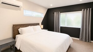 Een slaapkamer met een bed, nachtkastje, airconditioning, raam met gordijnen en een lamp.