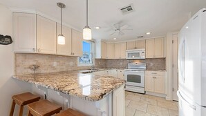 Eine Küche mit Granit-Arbeitsplatten, weißen Schränken, einem Edelstahl-Ofen, einer Mikrowelle und einem Kühlschrank. An der Kücheninsel stehen drei Holzbarhocker.
