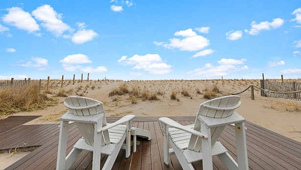 Blick auf einen Strand mit zwei weißen Adirondack-Stühlen auf einer Holzterrasse, einem sandigen Strand und einem Holzzaun im Hintergrund.
