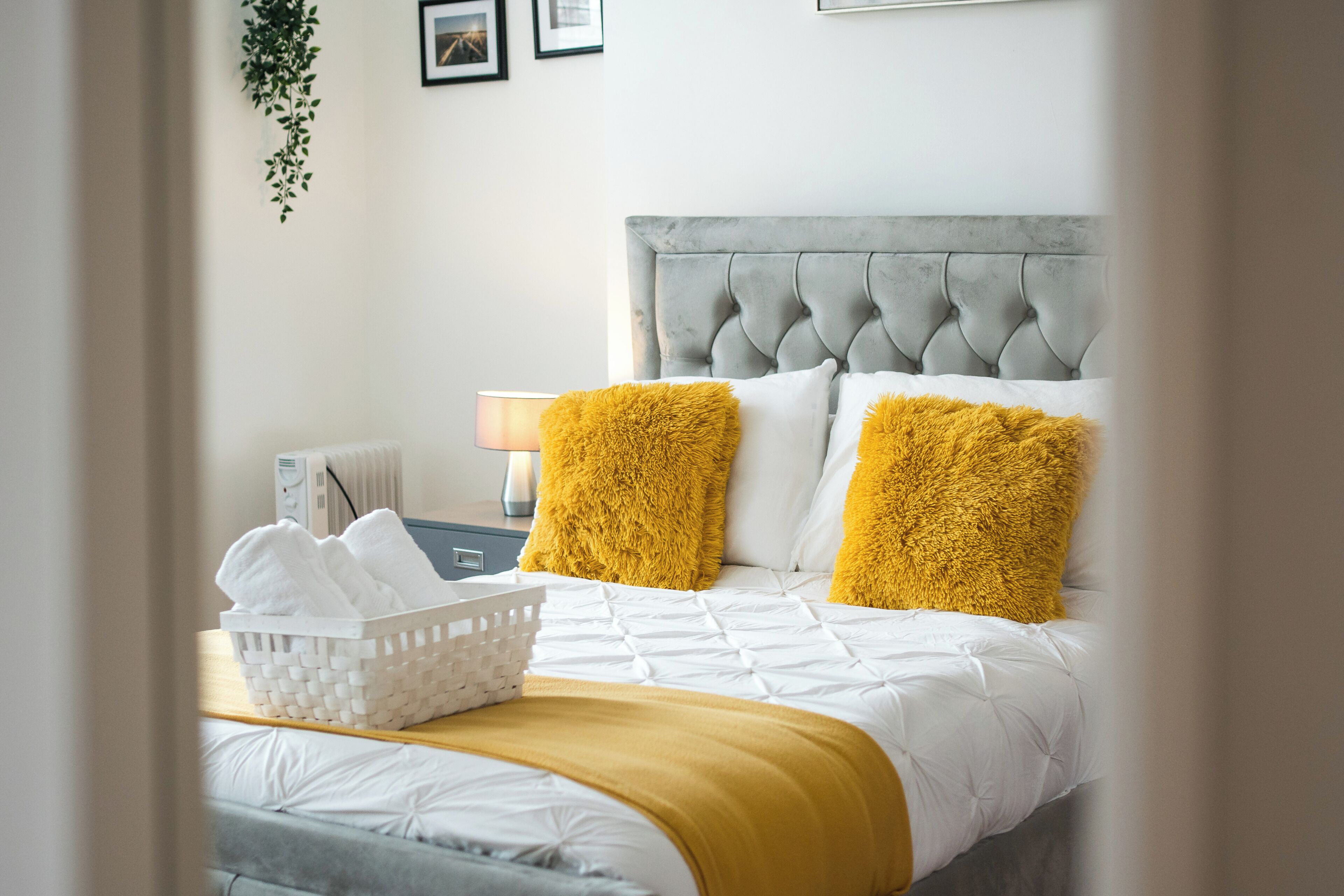 Una cama bien hecha con cabecera gris, ropa de cama blanca y dos almohadones amarillos. Hay una cesta blanca con toallas sobre la cama y una mesita de noche con una lámpara a la vista.