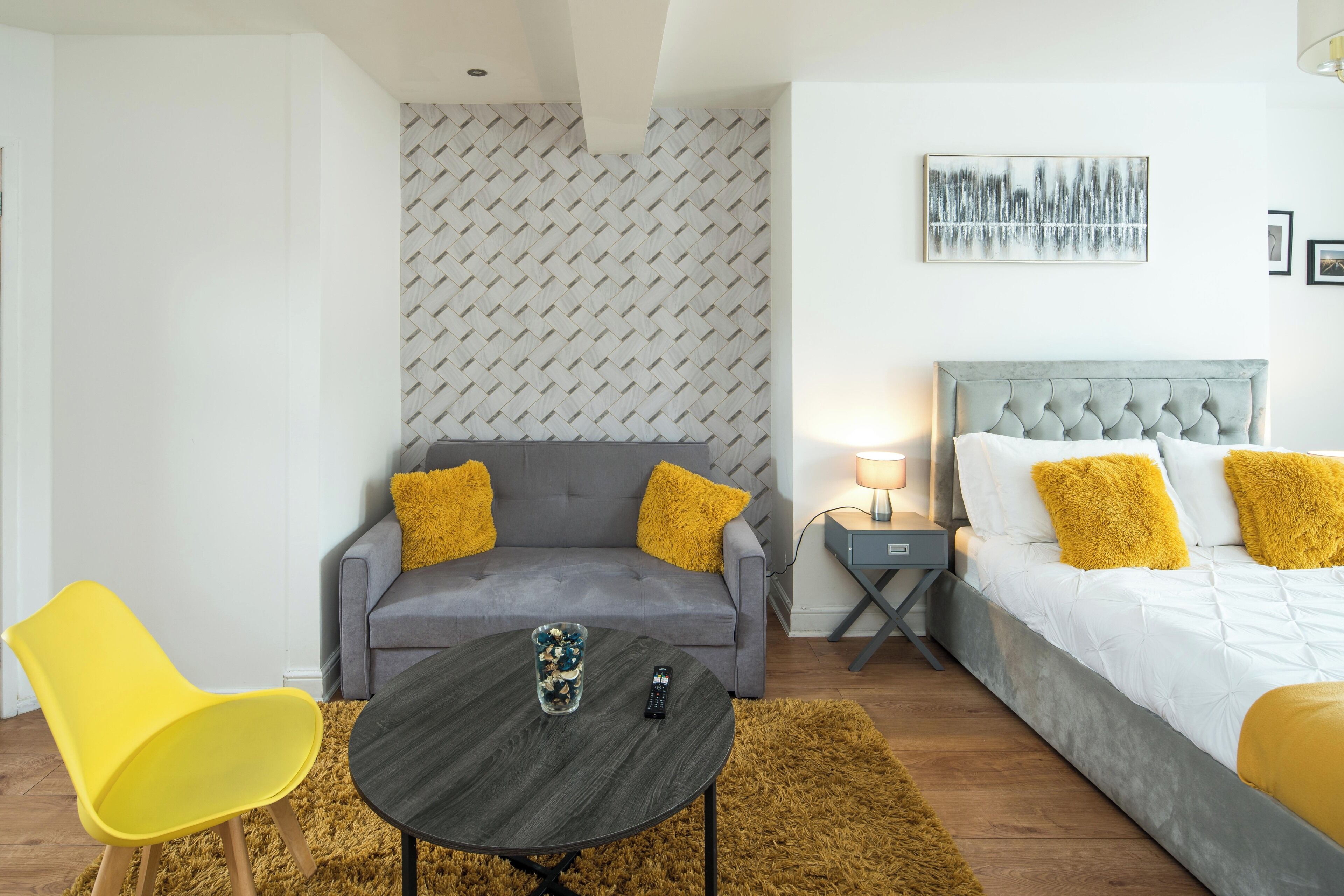 Une chambre d’hôtel moderne avec un canapé gris, une chaise jaune, un lit avec des oreillers blancs et jaunes, une tête de lit grise, une petite table avec une télécommande et une lampe fixée au mur.