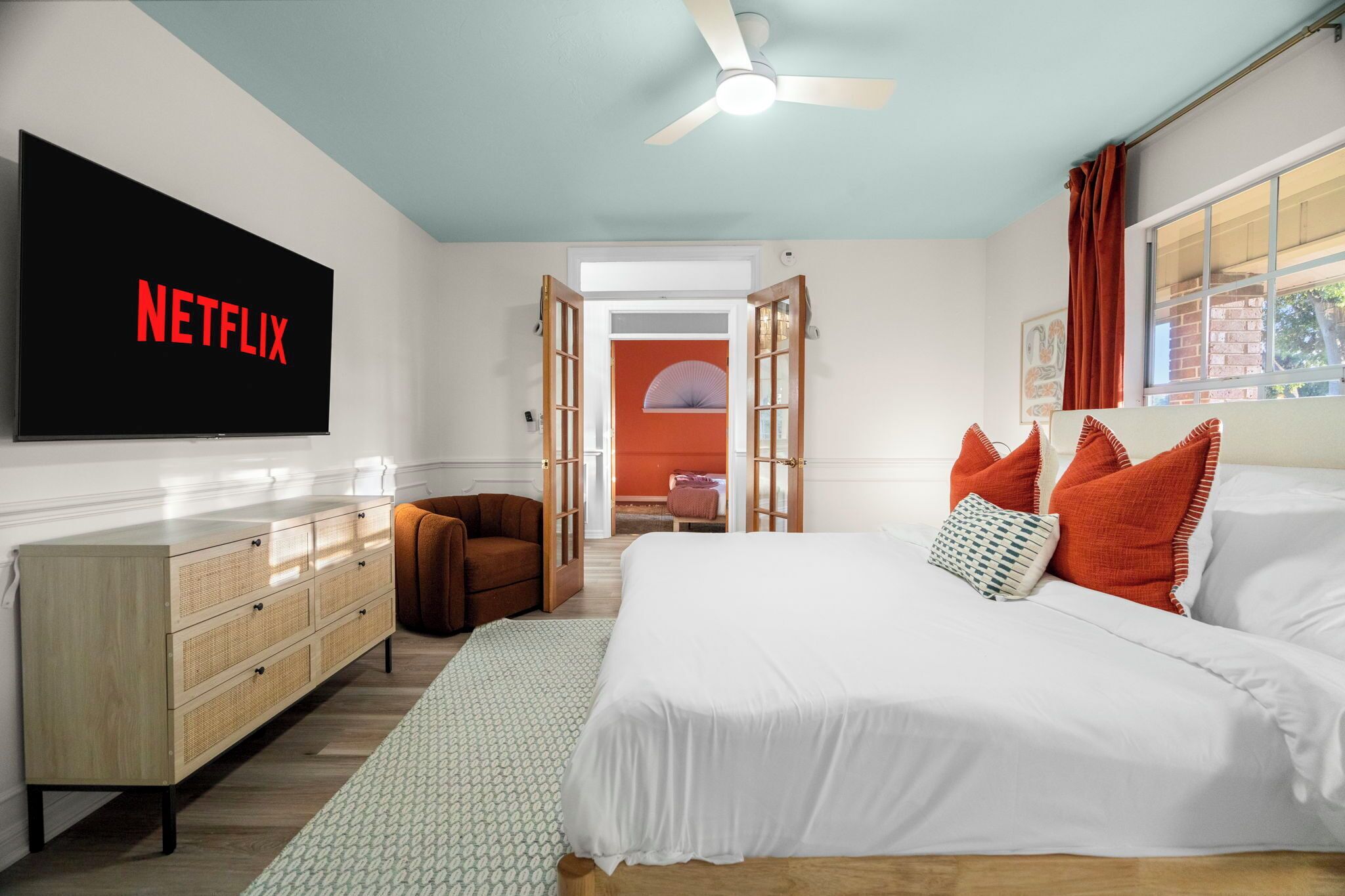 Ein Schlafzimmer mit einem Bett, einem Schminktisch, einem Sessel, einem Fernseher mit dem Schriftzug 'NETFLIX' und einem Deckenventilator.